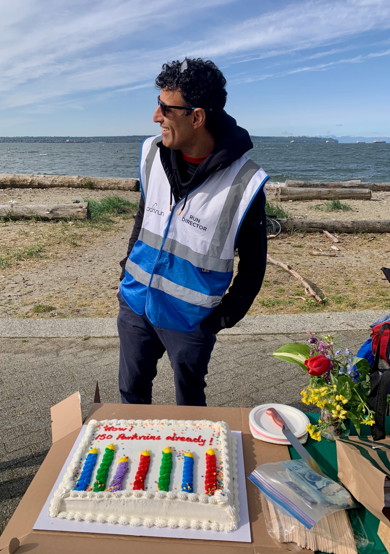 Jay Sidhu in a parkrun Run Director vest beside a cake marking 150 Ambleside parkruns